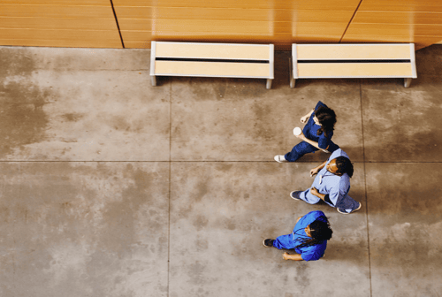 Aerial view of three people in business attire having a conversation while standing on a concrete floor in a modern building lobby with wooden benches visible in the background