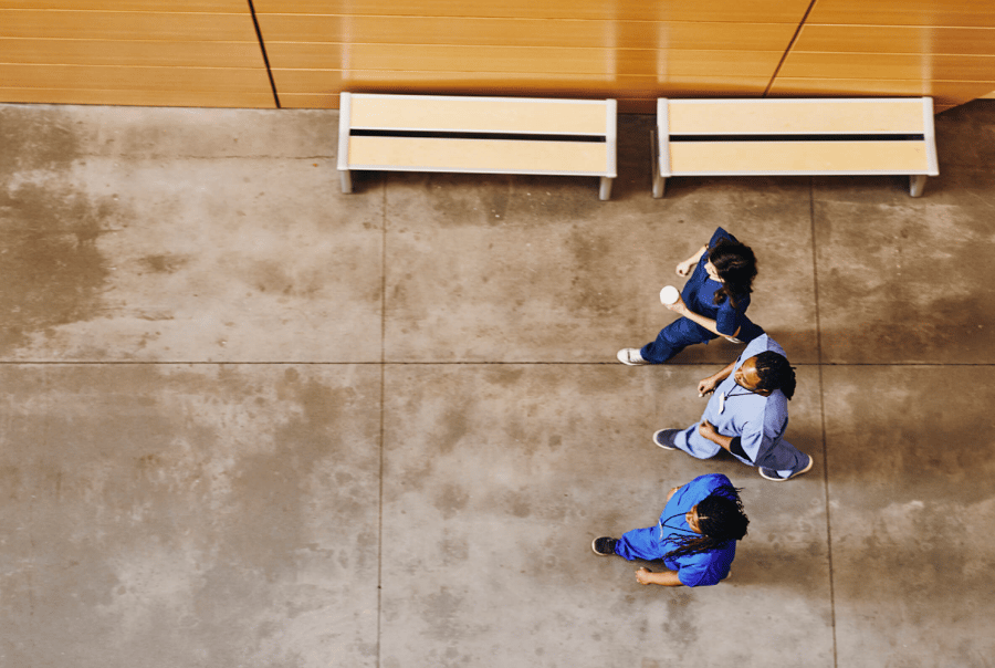 Aerial view of three people in business attire having a conversation while standing on a concrete floor in a modern building lobby with wooden benches visible in the background
