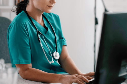 A healthcare professional in teal scrubs with a stethoscope works at a computer, shown from the shoulders down.