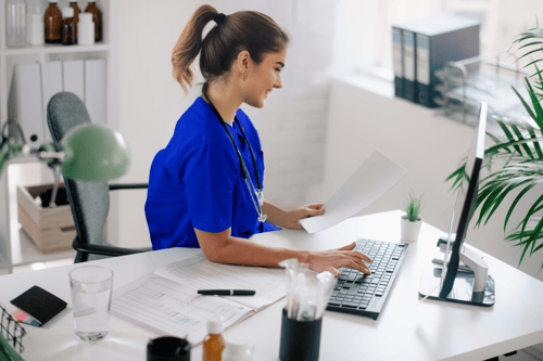 Young woman in bright blue medical scrubs with stethoscope smiling while typing on laptop computer at white desk in modern medical office, with papers, glass of water, and plant visible on workspace.