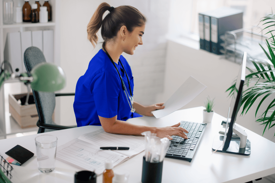 Young woman in bright blue medical scrubs with stethoscope smiling while typing on laptop computer at white desk in modern medical office, with papers, glass of water, and plant visible on workspace.