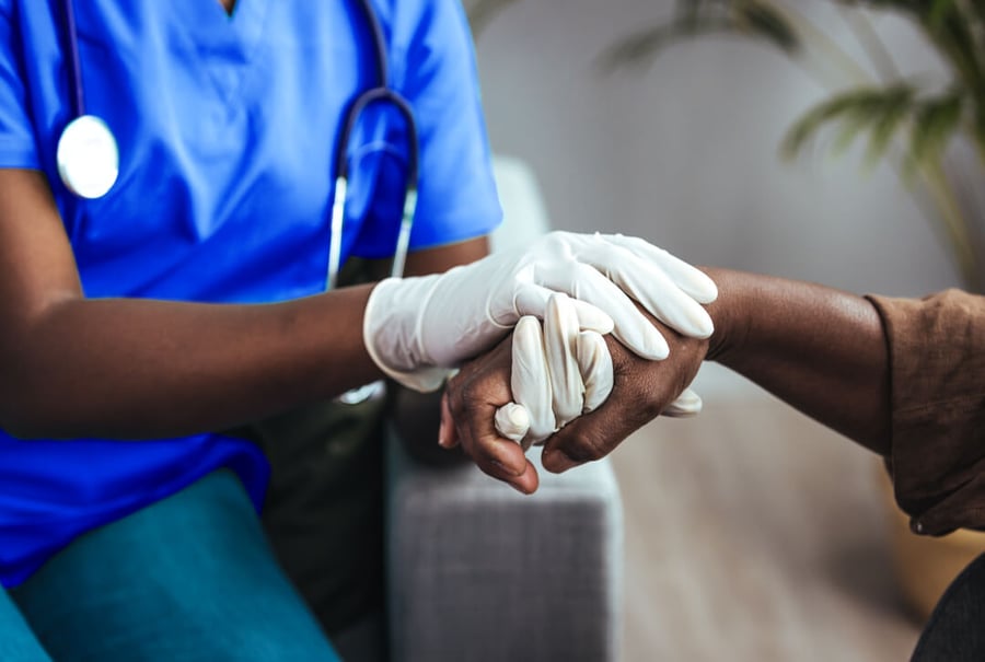A nurse in blue scrubs and white latex gloves gently holding a patient's hand, symbolizing compassionate healthcare and patient support
