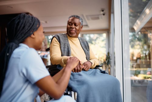 Caregiver compassionately holding hands with an elderly man in a wheelchair at a care facility