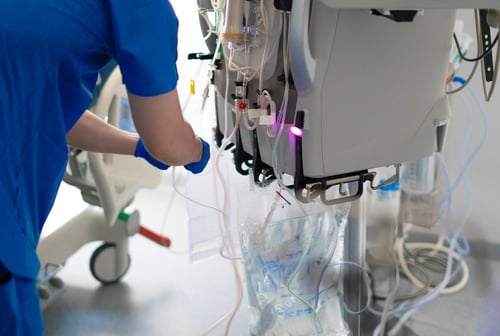 A healthcare professional in blue scrubs and gloves operating a dialysis machine with tubes and fluid bags in a clinical setting