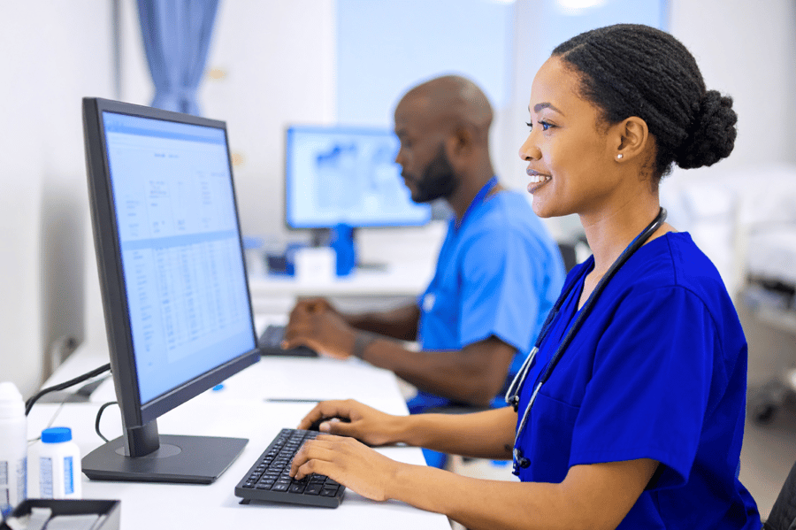 Female healthcare worker in blue scrubs with stethoscope typing on computer at medical workstation, with male colleague working in background in clinical setting