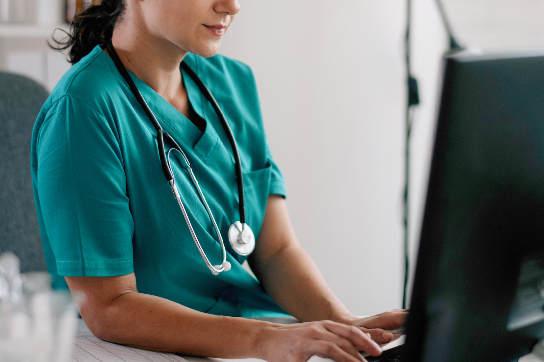 A healthcare professional in teal scrubs with a stethoscope works at a computer, shown from the shoulders down.