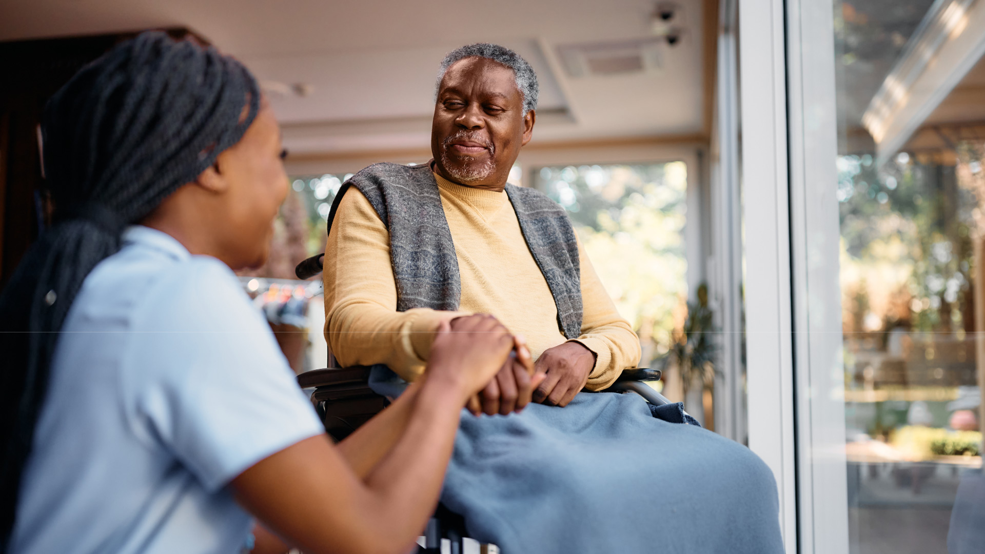 Caregiver compassionately holding hands with an elderly man in a wheelchair at a care facility