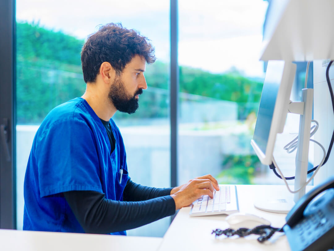 Clinician working at desktop computer in hospital office with large windows and natural lighting. Clinician working at desktop computer in hospital office with large windows and natural lighting.
