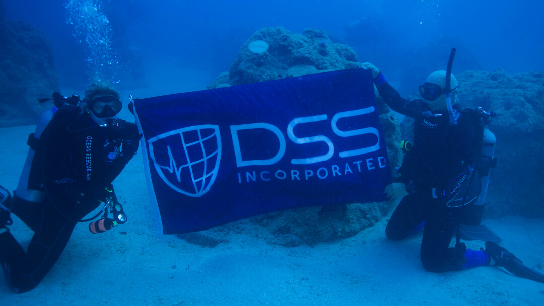 Two scuba divers underwater holding a blue banner with DSS Incorporated logo and company name on the ocean floor. Two scuba divers underwater holding a blue banner with DSS Incorporated logo and company name on the ocean floor.