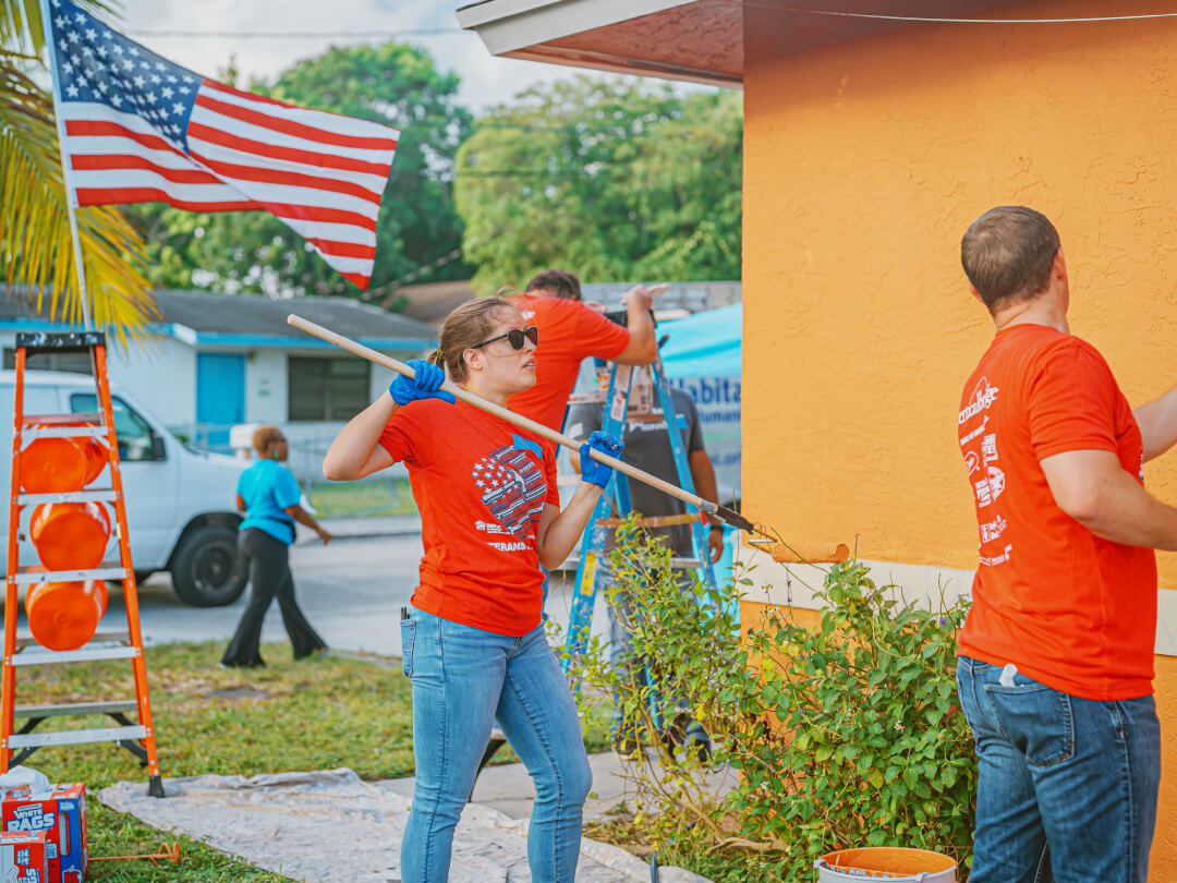Volunteers painting exterior walls of a house during a Habitat for Humanity community service project, with American flag visible in background. Volunteers painting exterior walls of a house during a Habitat for Humanity community service project, with American flag visible in background.