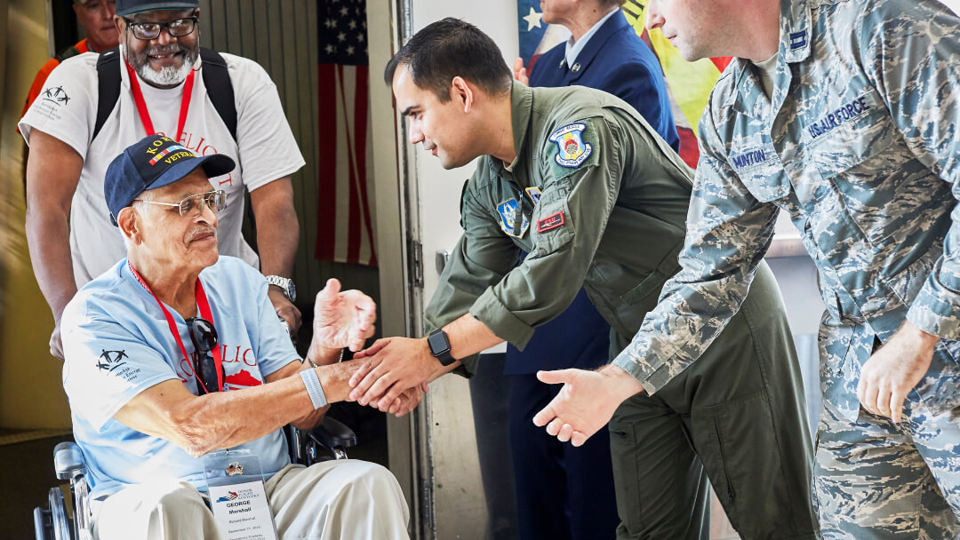 Military serviceman in flight suit shaking hands with elderly veteran in wheelchair wearing Vietnam War cap, surrounded by other veterans and active duty personnel at military ceremony or honor event. Military serviceman in flight suit shaking hands with elderly veteran in wheelchair wearing Vietnam War cap, surrounded by other veterans and active duty personnel at military ceremony or honor event.
