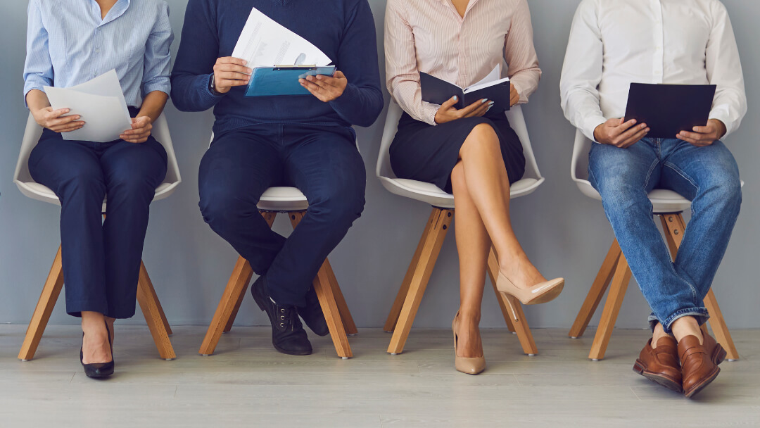 Four job candidates sitting in modern chairs holding resumes and documents while waiting for interviews in an office setting Four job candidates sitting in modern chairs holding resumes and documents while waiting for interviews in an office setting