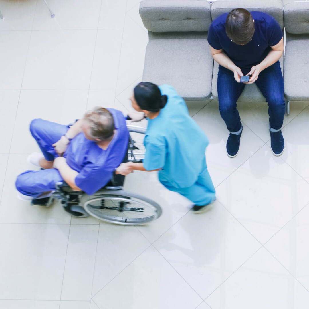 Medical professionals assisting a patient in a wheelchair in a hospital waiting area, with another person sitting on a couch in the background Medical professionals assisting a patient in a wheelchair in a hospital waiting area, with another person sitting on a couch in the background