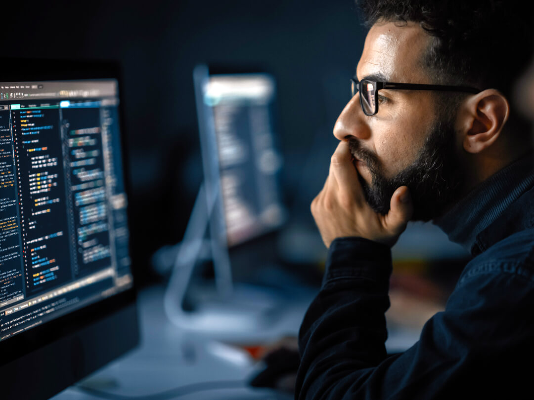 Software developer with glasses and beard working late at night, looking thoughtfully at computer screen displaying code in a dimly lit office