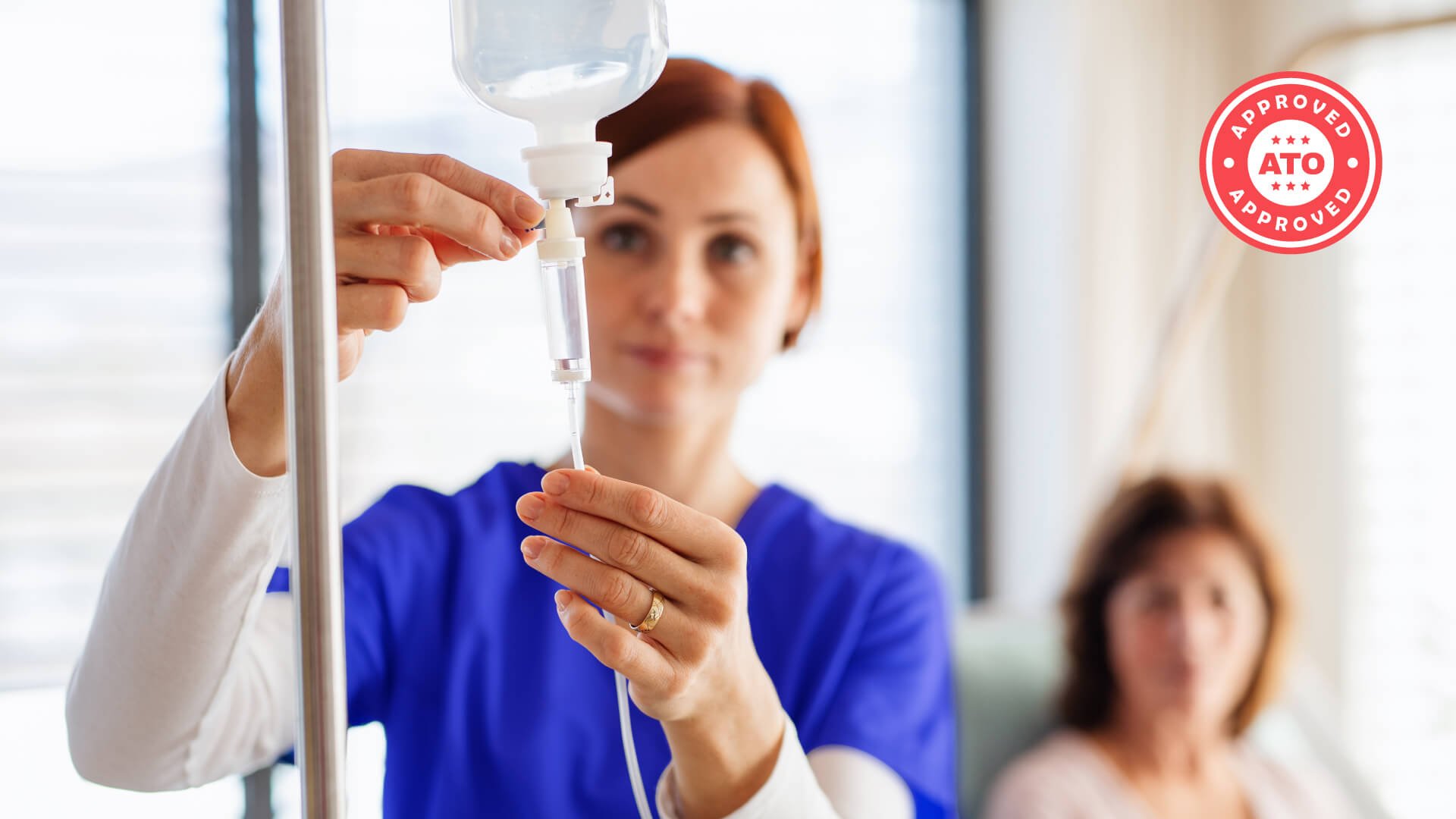 Healthcare professional in blue scrubs adjusting an IV drip line in a medical setting with patient in background