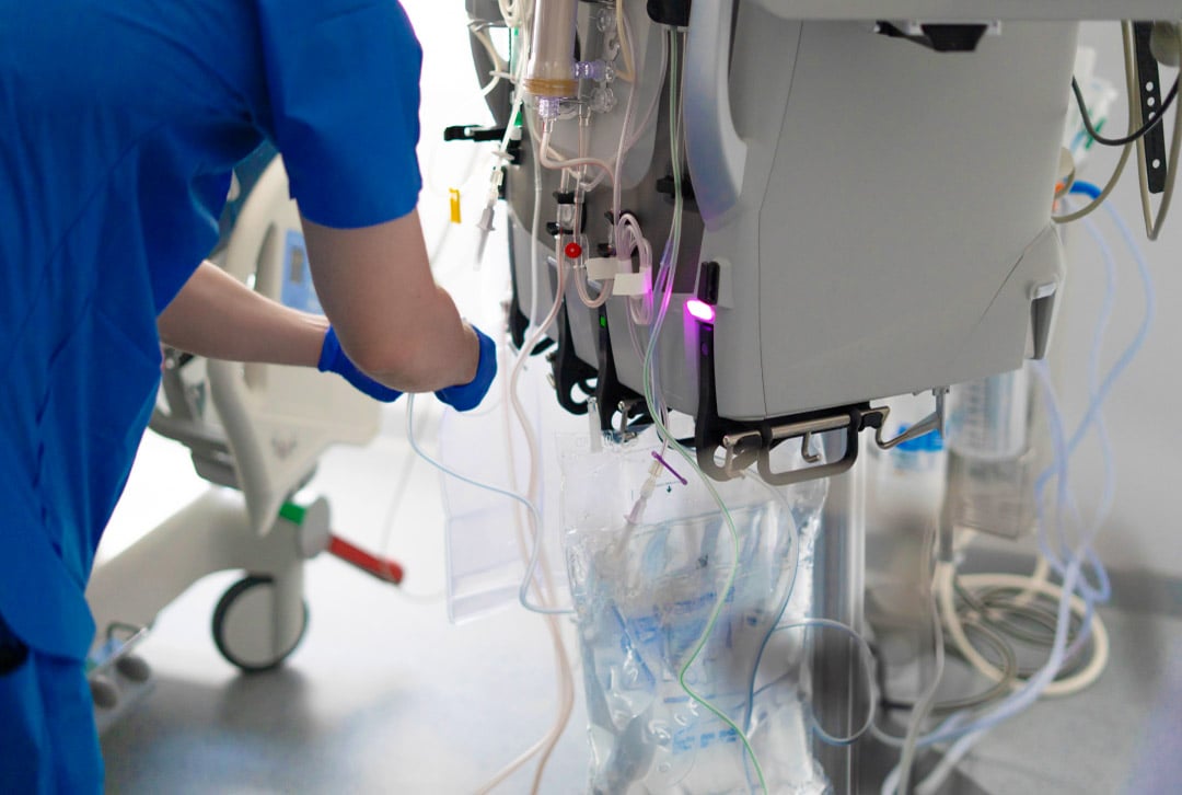 A healthcare professional in blue scrubs and gloves operating a dialysis machine with tubes and fluid bags in a clinical setting