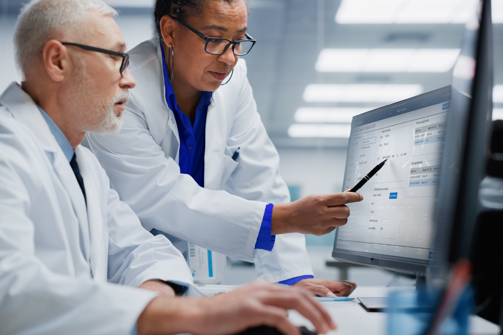 Two doctors in lab coats review data on a computer screen, one seated and one standing, collaborating in a clinical environment.