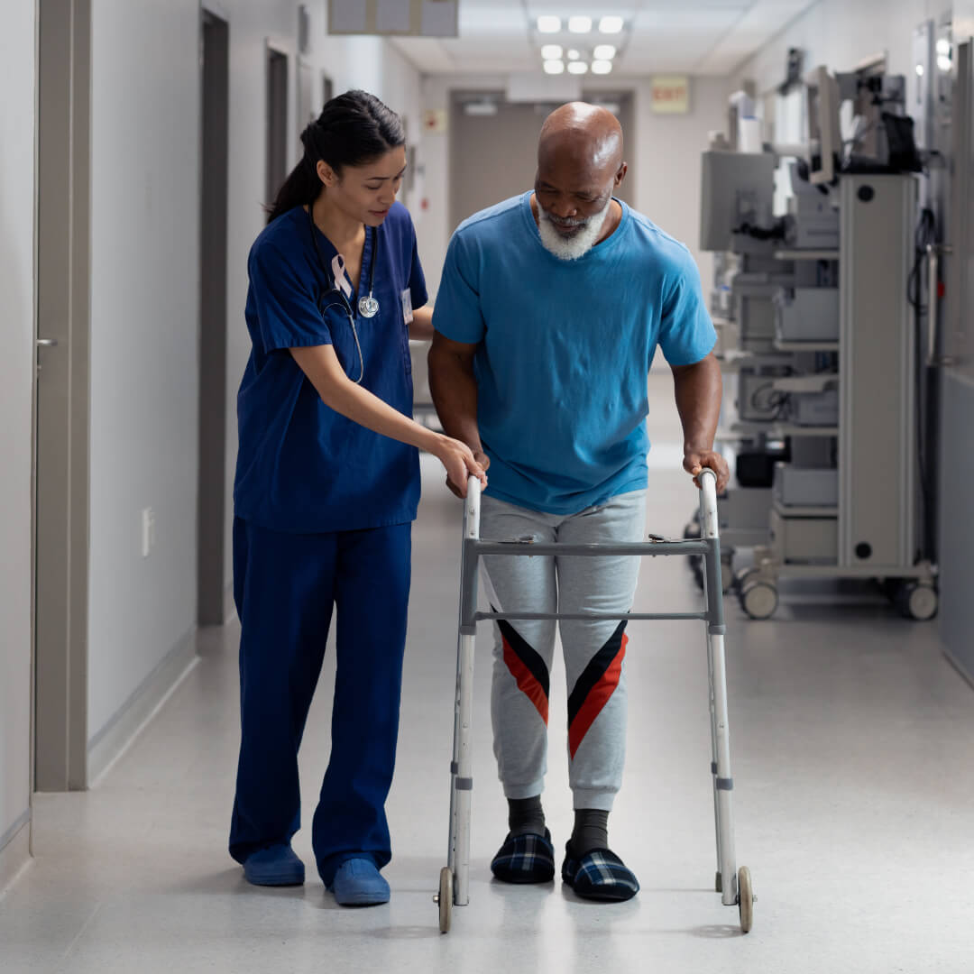 Healthcare worker in blue scrubs assisting elderly patient with walker in hospital hallway during physical therapy rehabilitation